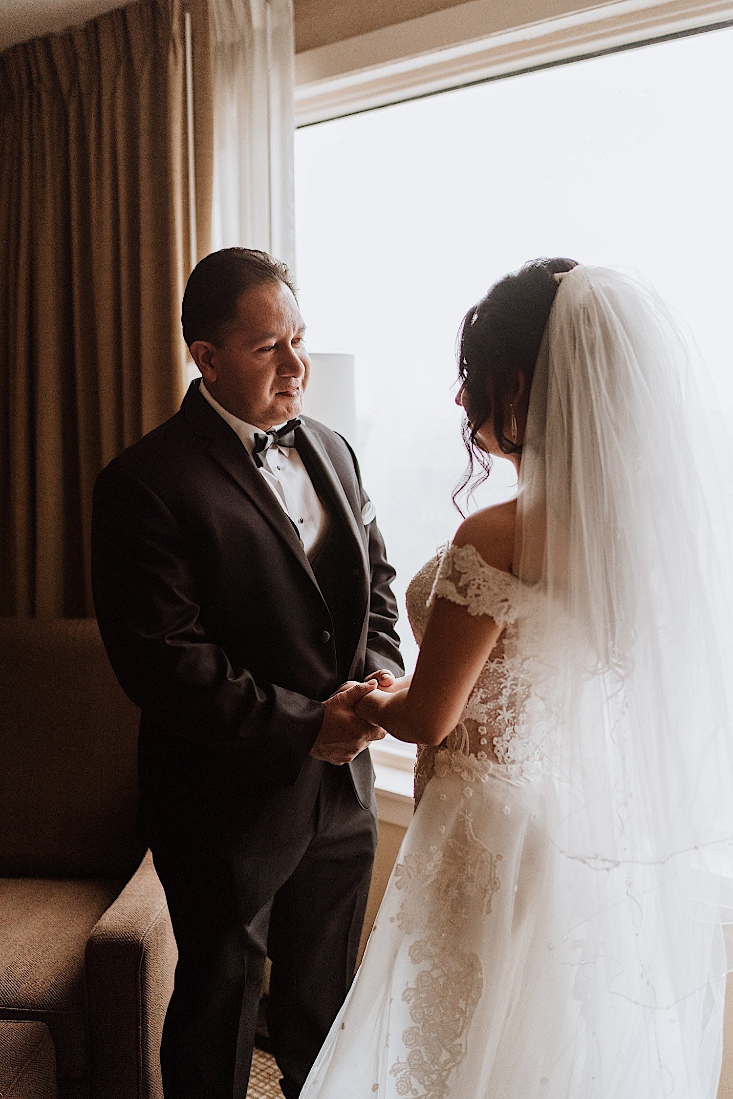Father holds hands with his daughter in her wedding dress in front of a window