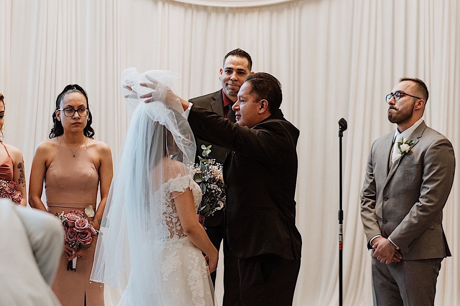 Father lifts the veil over his daughters head as she prepares for her wedding ceremony as her soon to be husband looks on