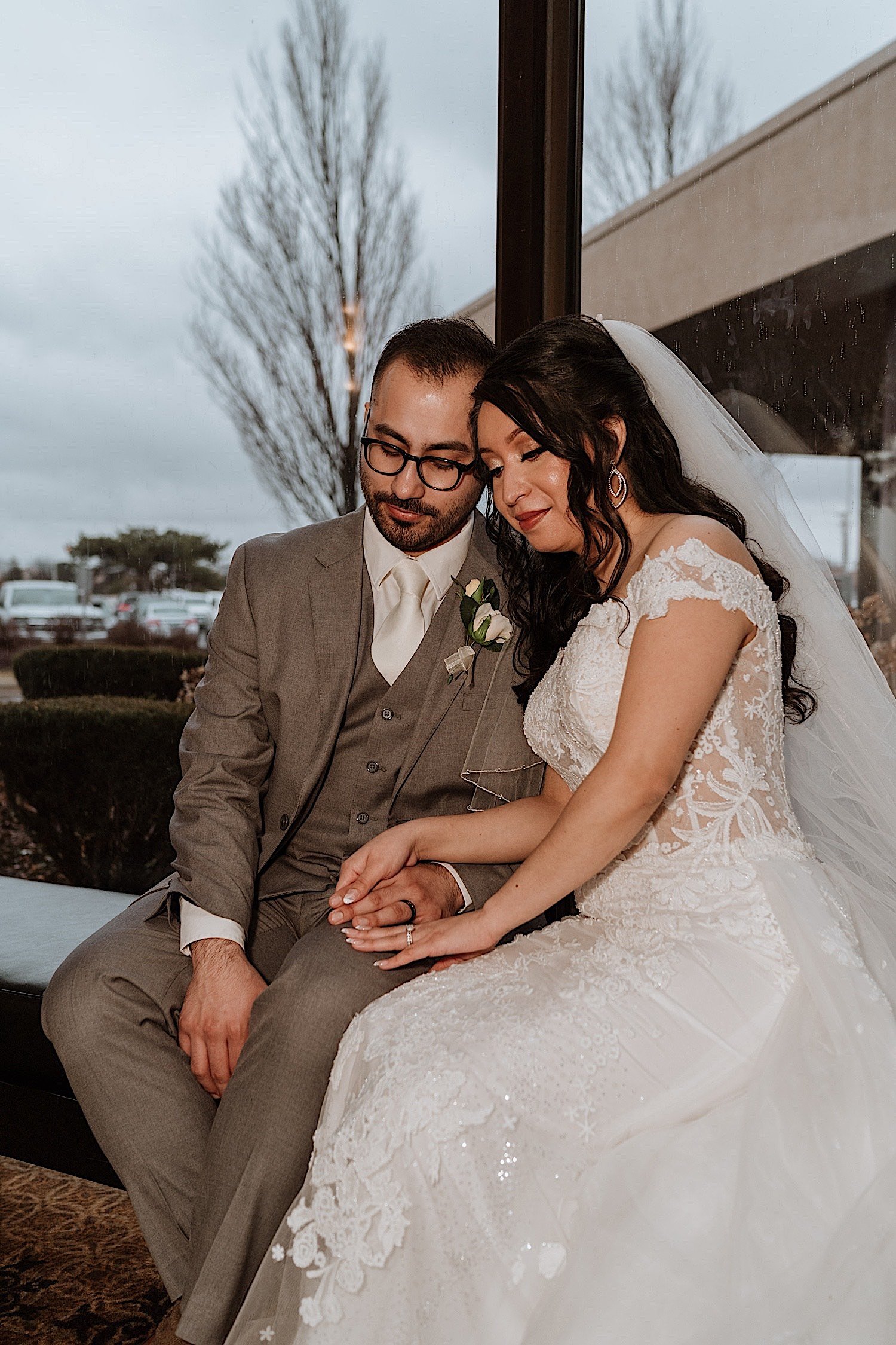 Bride and groom seated with each other looking at their wedding rings together with a window behind them