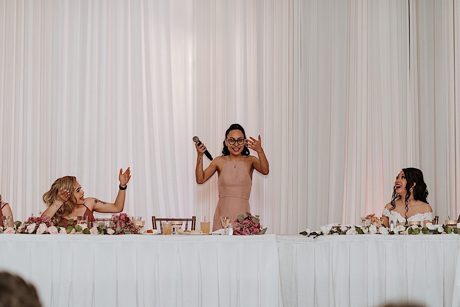 Bridesmaid stands up to give a speech at wedding reception as bride laughs next to her