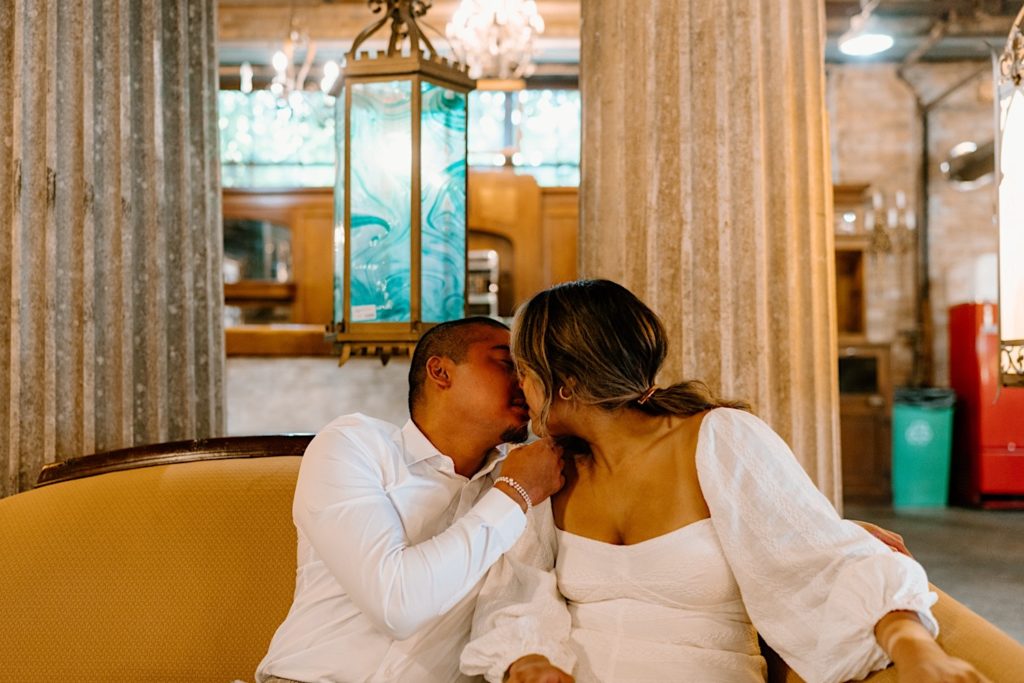 An engaged couple sits on a yellow vintage couch almost kissing and wearing all white during their engagement session at Salvage One in Chicago.