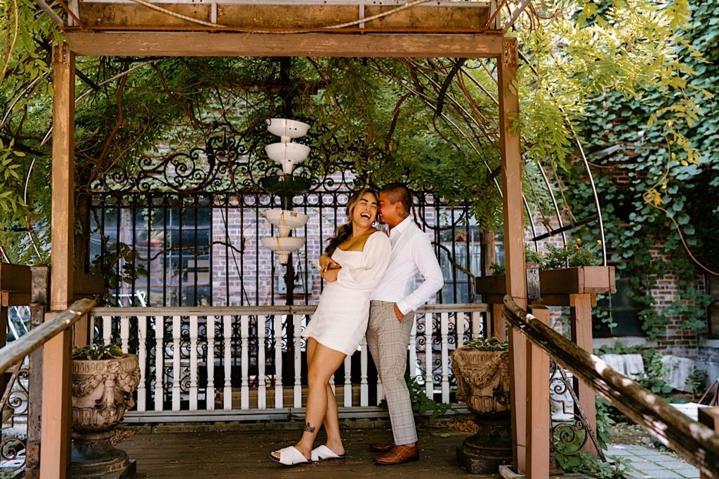 A couple stands under a pergola at Salvage One in Chicago smiling and laughing at one another during their engagement session.
