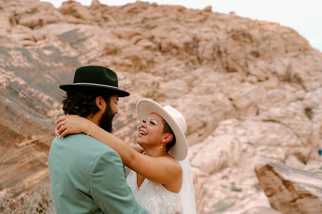 A bride and groom hold one another in front of the Red Rocks, the bride is wearing a two piece white dress and wide brimmed hat with a veil.  The groom his wearing a green suit.