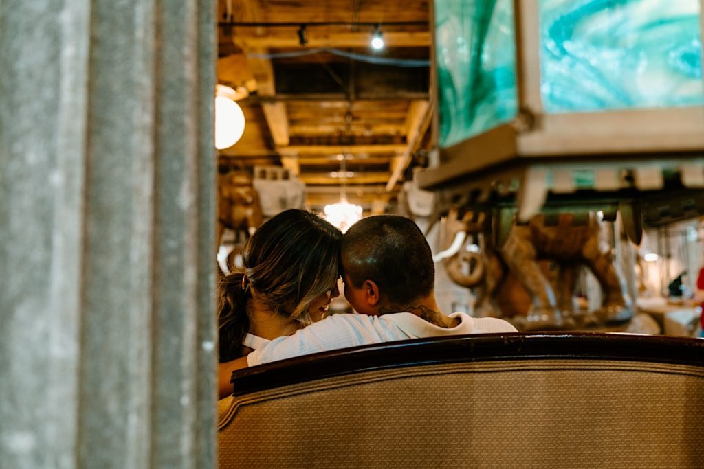 A couple sits together on a vintage couch with their foreheads pressed together during their engagement session.  There is a blue vintage stained glass light in the foreground of the image. 