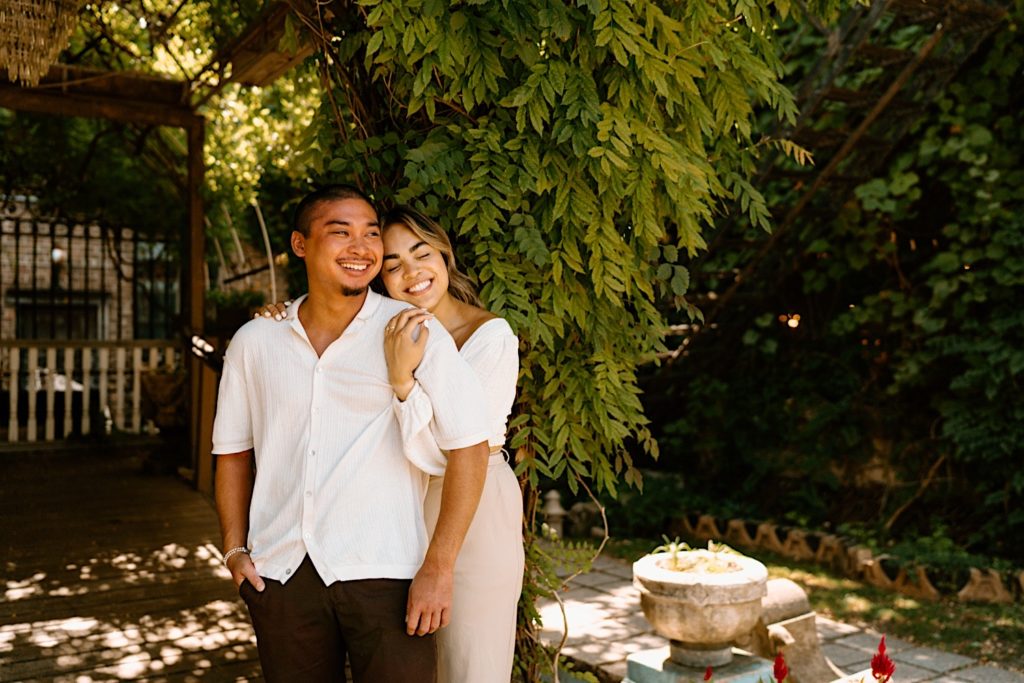 A fiancée holds her fiance close, with his back to her chest.  They are standing outside and smiling at Salvage one in Chicago.