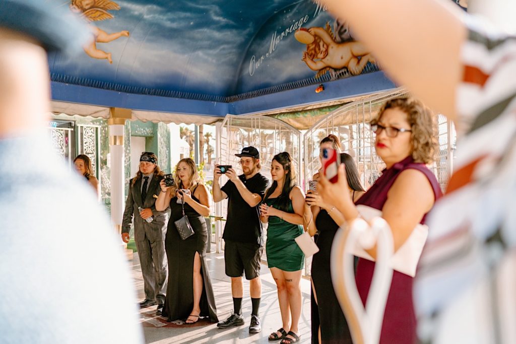 Wedding guests record the ceremony with their phones while the couple sits in the pink convertible at the White Little Chapel.