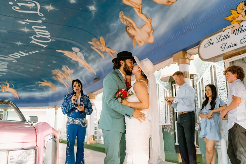 The bride and groom kiss in the pink convertible while Elvis oversees the last of their wedding ceremony.