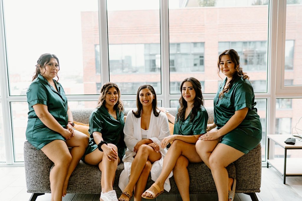 The bride and her bridal party sit smiling at the camera in matching pajamas.  The bride wears white while the bridesmaids wear green sating pajamas.