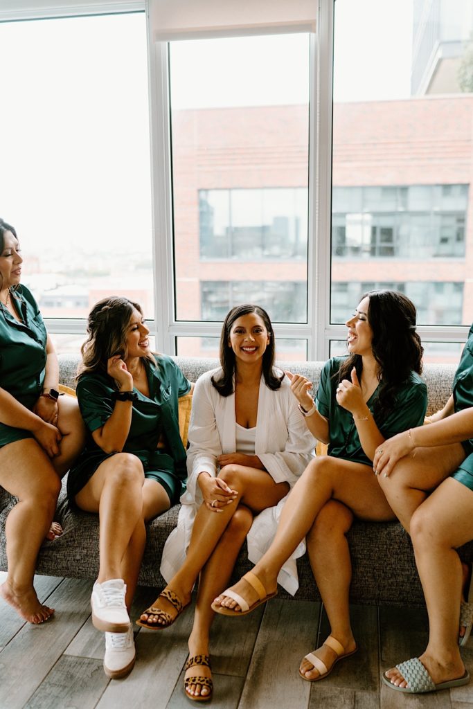 The bride and her bridal party sit smiling at the camera in matching pajamas.  The bride wears white while the bridesmaids wear green sating pajamas.