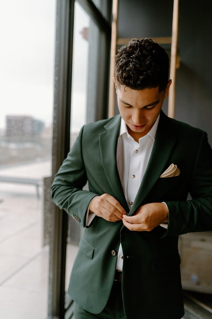 The groom stands with his green suit in front of a window on his wedding day in his Chicago Airbnb