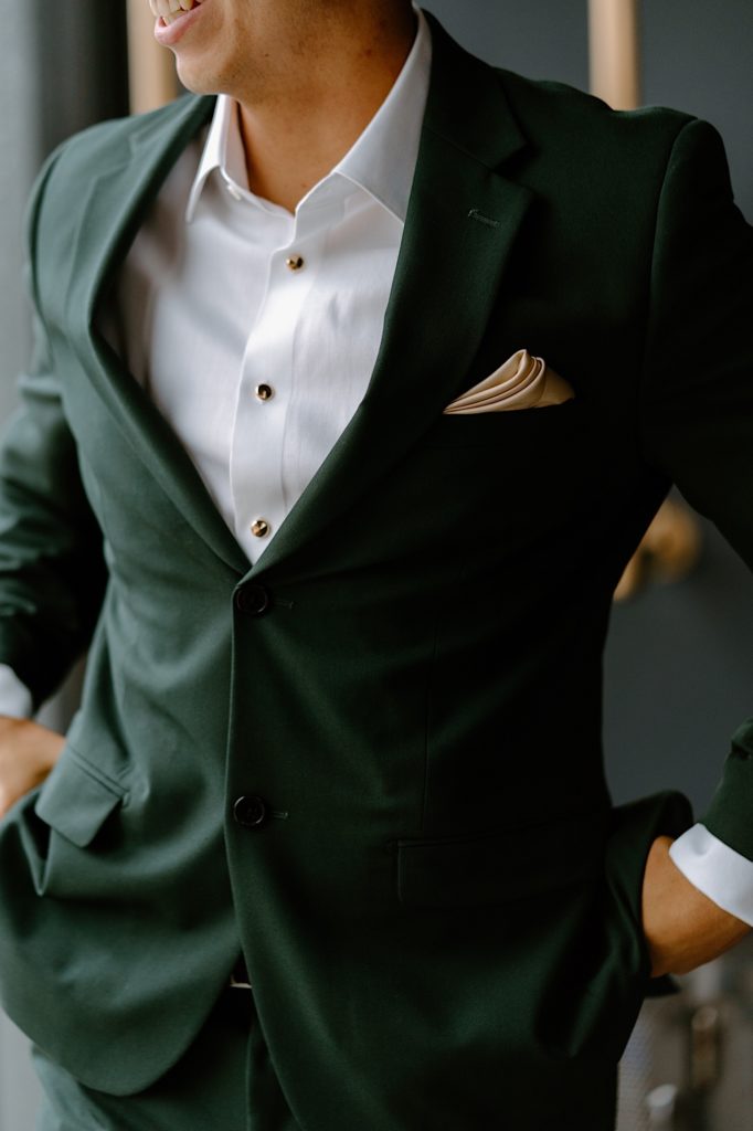 The groom stands with his green suit in front of a window on his wedding day in his Chicago Airbnb