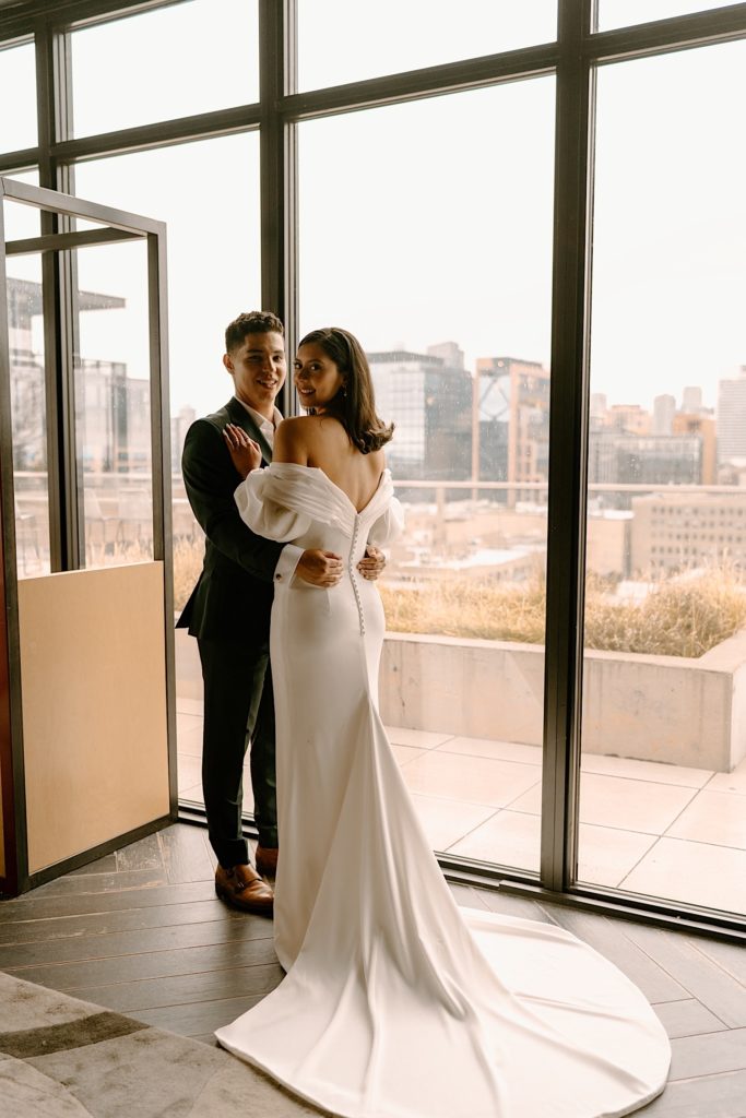 A bride and groom hold one another in front of a window overlooking the city before their wedding at Loft Lucia.