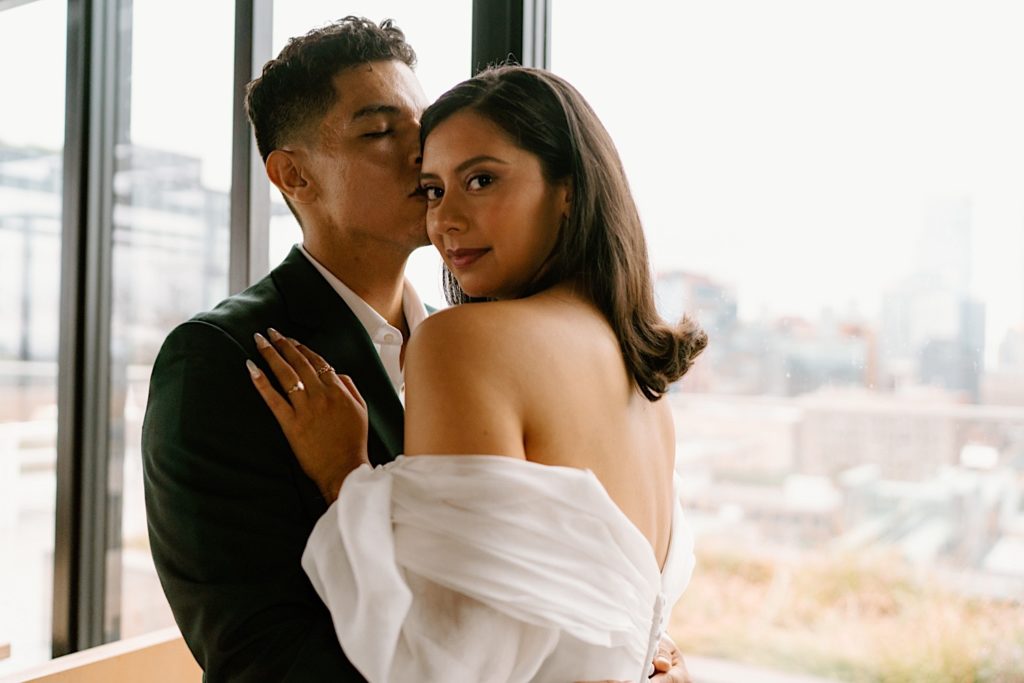 A bride and groom hold one another in front of a window overlooking the city before their wedding at Loft Lucia.