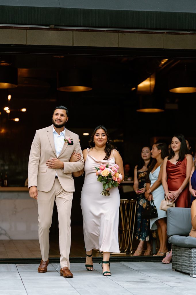 The wedding party in beige suits and pink dresses walk down the aisle to the wedding ceremony on a Chicago rooftop.