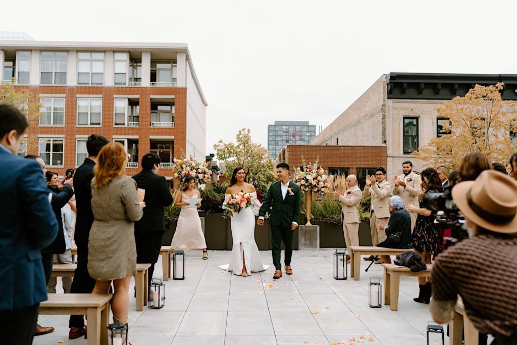 The bride and groom hold hands leaving their ceremony space on a rooftop at Loft Lucia in Chicago.