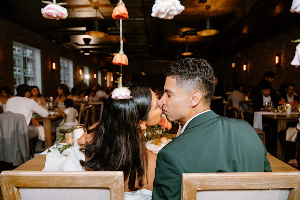 The newlyweds kiss at their sweetheart table decorated with dangling flowers at their reception at Loft Lucia