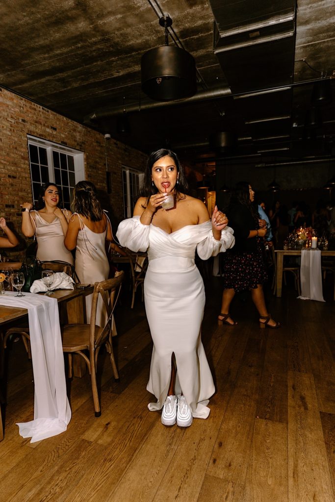 The bride stands sipping a drink during her wedding reception with cute sneakers on.