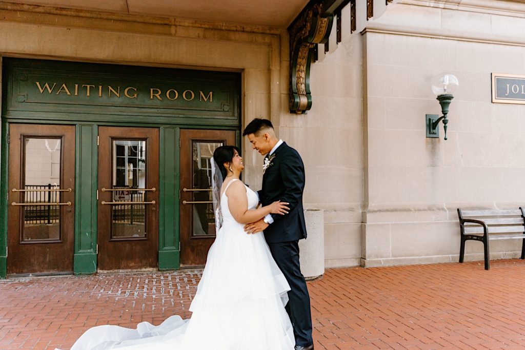 A bride and groom stand holding one another during their first look standing underneath the overhand at Joliet's Union Station.