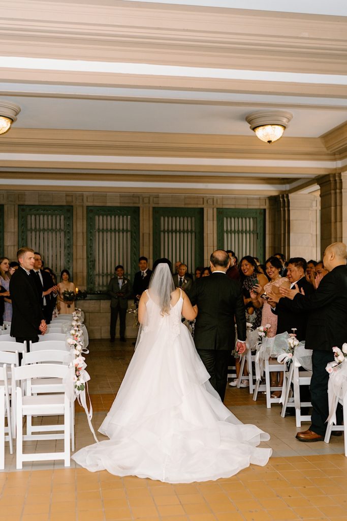 The bride and her father walk down the aisle to her wedding ceremony framed by greenery, chairs and lit candles.