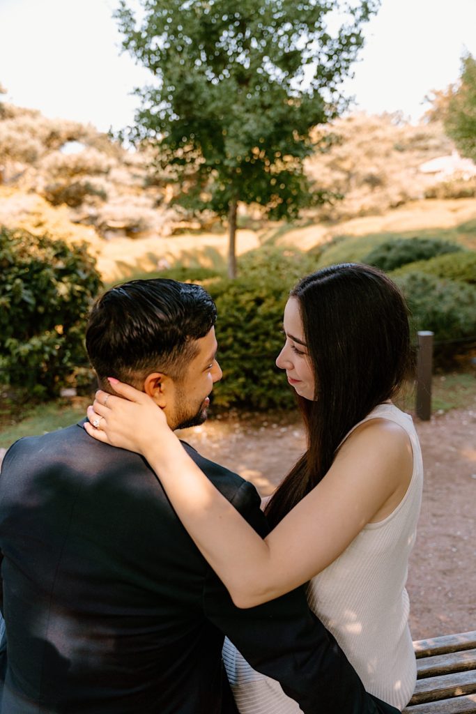 A bride rests her hand on the neck of her fiancé during their engagement session.  You can see her ornate engagement ring. 