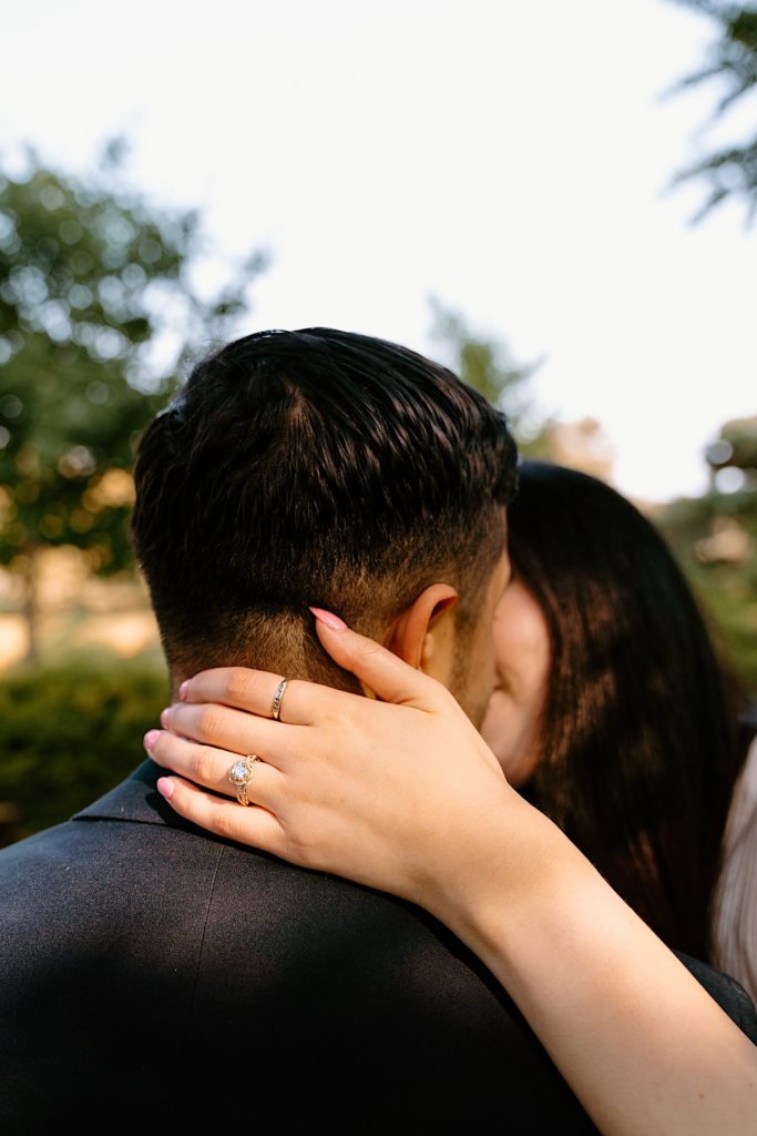 A bride rests her hand on the neck of her fiancé during their engagement session.  You can see her ornate engagement ring. 