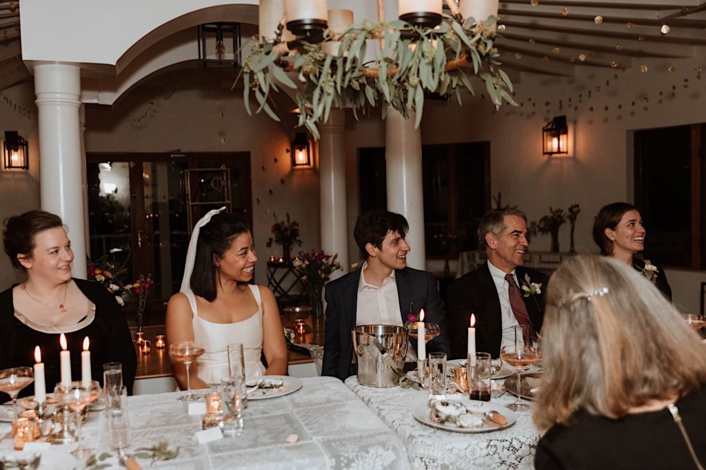 A bride and groom smile at their guests as they give a speech on their wedding day at their Airbnb wedding venue.