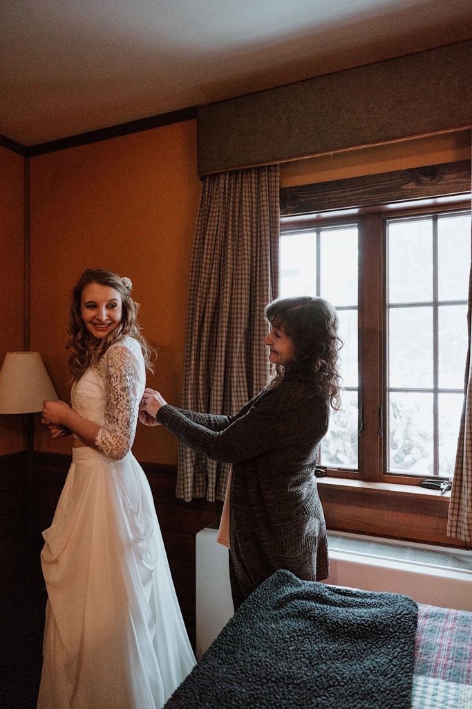 A bride stands while her mother buttons the back of her dress.  She looks back at her mom smiling.  