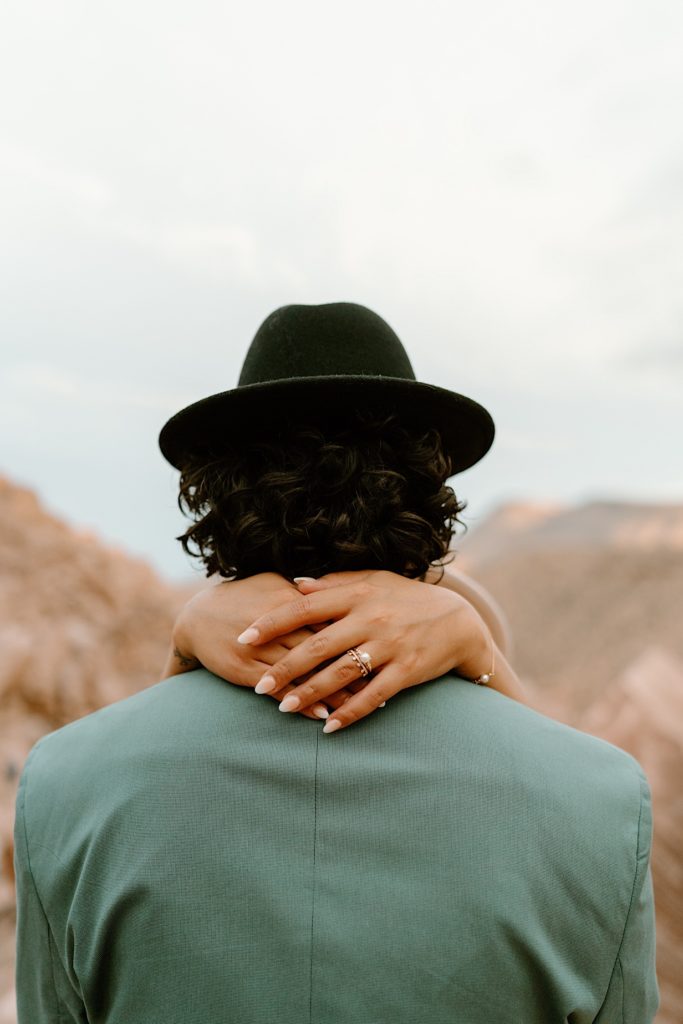 A groom stands with his back to the camera while his bride holds her hands around his neck.