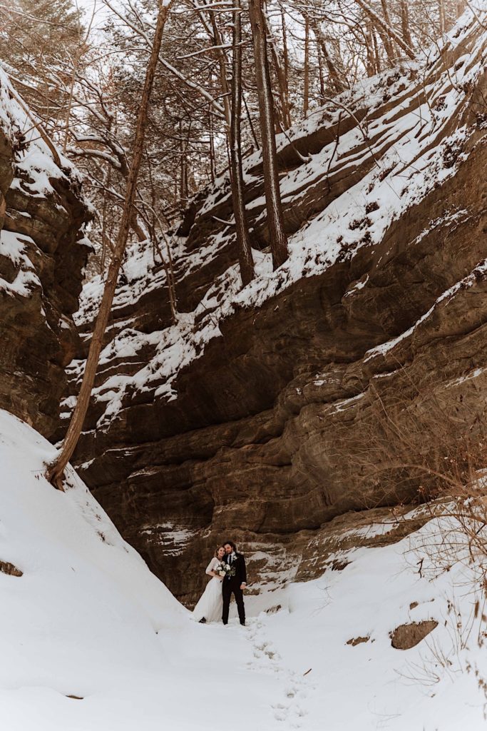 A bride and groom stand in the snow next to a cliff while smiling at the camera.