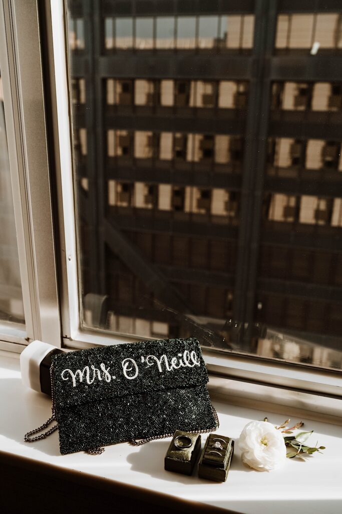 A bridal purse with the brides future name written in glitter on the purse.  The purse sits with other details from the brides wedding day including flowers and the wedding rings.  