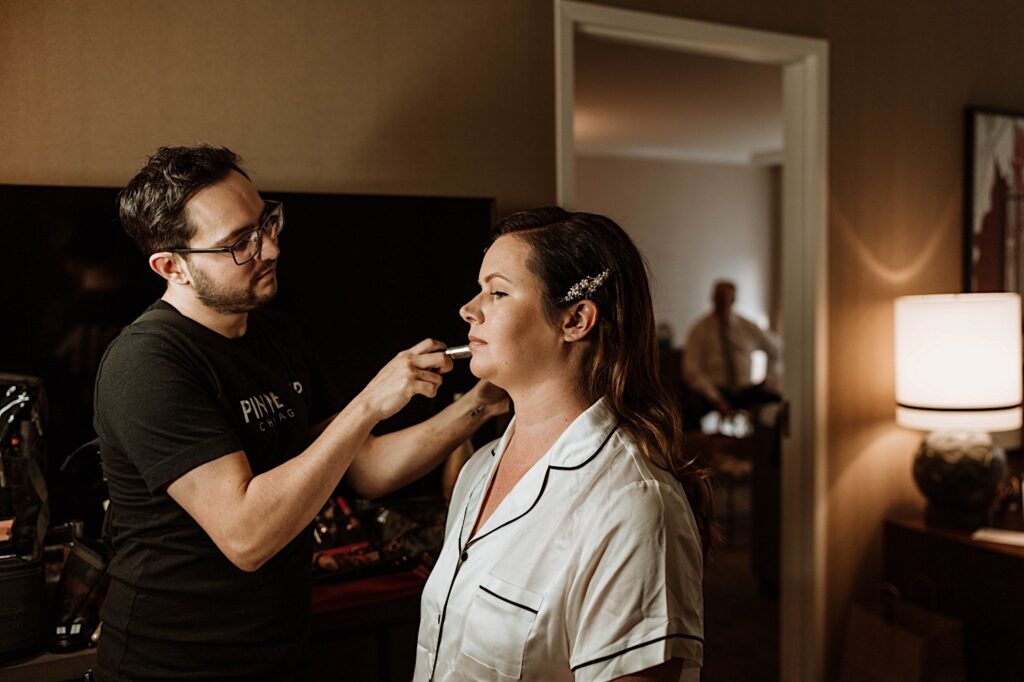 The bride sits in cream satin bridal pajamas having her makeup applied by her hair and makeup artist from Pin Me Up Chicago