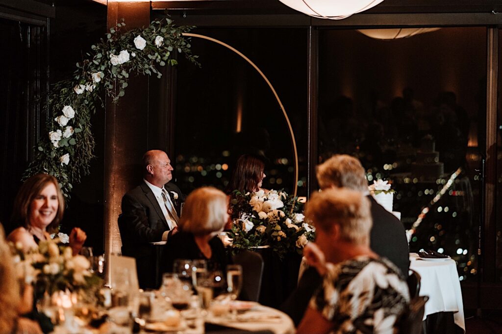 A bride and groom smile and laugh at a speech given by a guest during their wedding reception on the 95th floor.