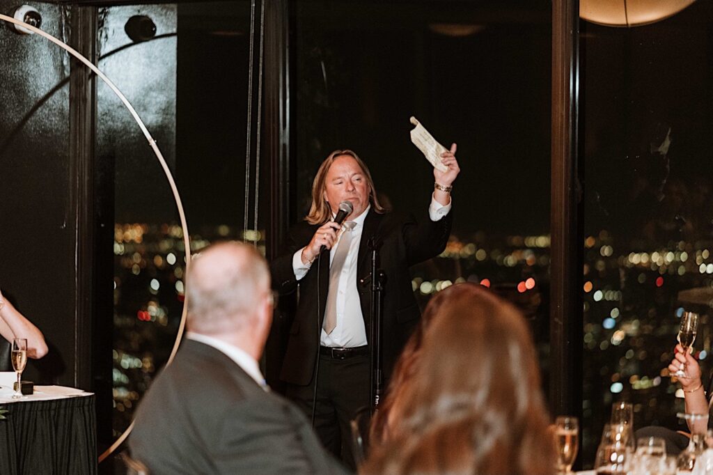A guest gives a speech at a wedding in front of a window overlooking the city of Chicago