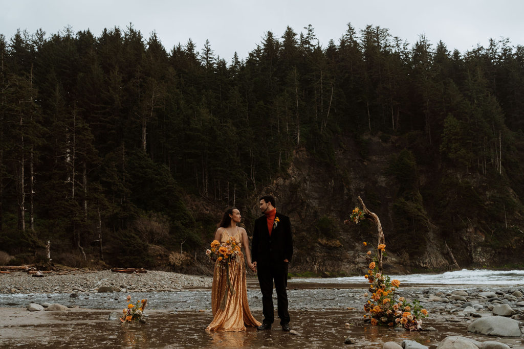 A couple holds hands while looking at one another during their elopement ceremony.  They are surrounded by orange flowers during their elopement ceremony.