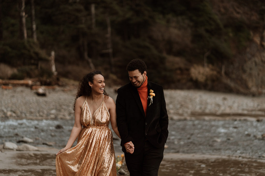 A bride and groom walk together holding hands after eloping on the beach.  