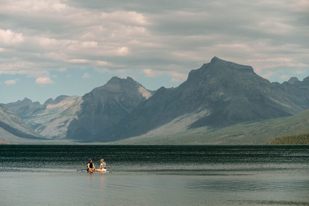 A couple sitting on a paddle board in a lake in front of mountains at Glacier National Park.