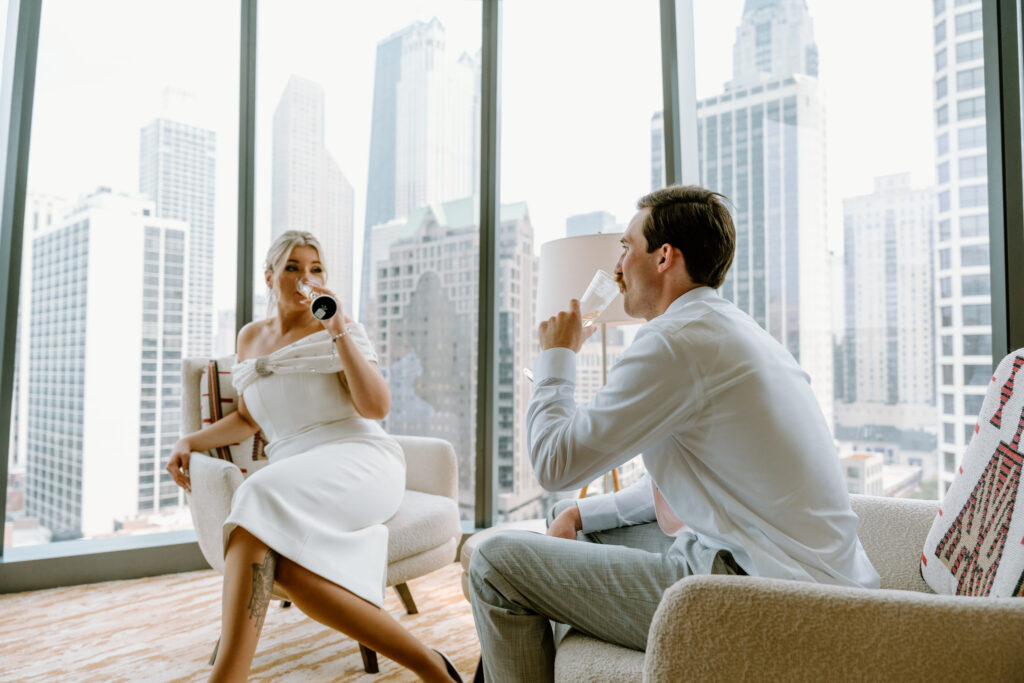 bride and groom drinking champagne in their hotel room with a floor to ceiling backdrop of the chicago skyline